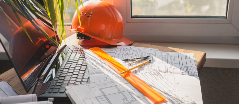 Construction workspace with detailed blueprints, a bright orange hard hat, a laptop, and drafting tools, emphasizing precision in construction documentation.